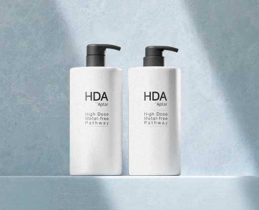 Two white cylindrical pump bottles with black dispensers labeled “HDA by Aptar High Dose Metal-free Pathway” showcase a high dose dispensing pump design, standing side by side on a light blue surface against a textured blue background.
