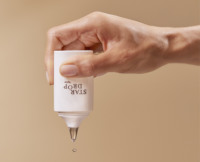 A hand is holding a white bottle labeled STAR DROP upside down, using drop-by-drop dispensing to release a single drop of liquid from the applicator against a plain beige background.