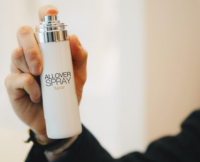 A hand holds a white bottle with silver actuator labeled ALLOVER SPRAY by Aptar, showcasing premium aerosol packaging labeled All Over Spray against a softly blurred, light-colored background.