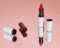 Two silver and black refillable lipstick packaging tubes with bright red tips are placed on a pink reflective surface. One lipstick stands upright with the cap removed
