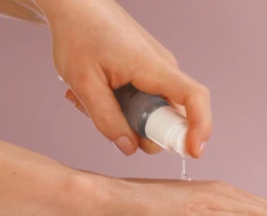 A hand dispenses clear liquid from an airless packaging pump bottle onto the back of another hand against a soft pink background.