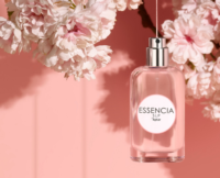 A clear perfume bottle with a low-profile fragrance pump labeled ESSENCIA SLP by Aptar hangs beside blooming pink and white flowers against a soft pink background.