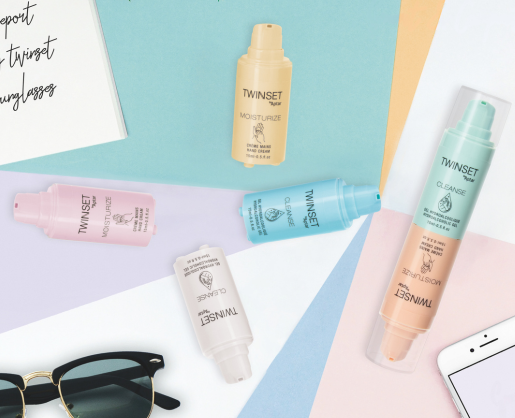 Five colorful TWINSET skincare bottles with innovative 2-in-1 Airless Packaging are arranged on pastel paper backgrounds near sunglasses, a smartphone, and a handwritten notepad. Each bottle is labeled for a different skincare step, like moisturize or cleanse.