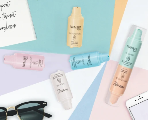 Five colorful TWINSET skincare bottles with innovative 2-in-1 Airless Packaging are arranged on pastel paper backgrounds near sunglasses, a smartphone, and a handwritten notepad. Each bottle is labeled for a different skincare step, like moisturize or cleanse.