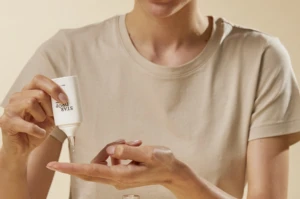 A person in a beige t-shirt squeezes lotion from a small white tottle named Star Drop onto their fingertips, preparing to apply it to their skin.