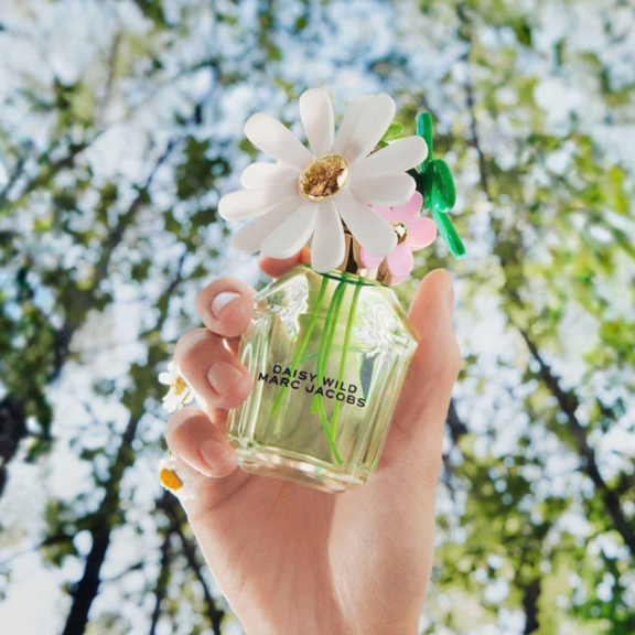A hand holds a bottle of Marc Jacobs Daisy Wild perfume, featuring VP4, a High-Performing Fragrance Pump labeled and a flower-shaped cap, set against tall, blurred green trees and a radiant sky.