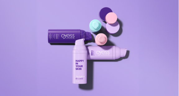 Three purple bottles of Aptar Beauty's best-selling airless packaging named Mezzo are shown in the market from the brand Oyess. Pastel-colored lids are arranged on a purple surface. One bottle reads HAPPY IN YOUR SKIN and Live kind to the world. The background and caps are pastel shades.