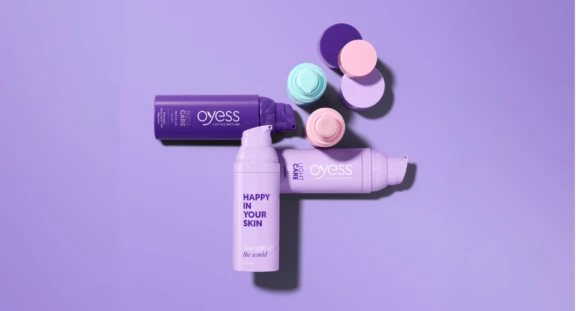 Three purple bottles of Aptar Beauty's best-selling airless packaging named Mezzo are shown in the market from the brand Oyess. Pastel-colored lids are arranged on a purple surface. One bottle reads HAPPY IN YOUR SKIN and Live kind to the world. The background and caps are pastel shades.
