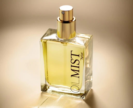 Clear glass bottle with a gold spray pump for dry oils, filled with yellow liquid and labeled 'OIL MIST by Aptar' in black text, shown against a softly lit beige background.