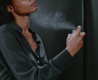 A person in a dark, silky shirt uses a prestige fragrance pump to spray perfume on their neck, creating a visible mist against a dark background. The image is cropped to show their lower face, neck, and hand holding the bottle.