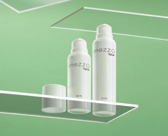 Two white plastic bottles labeled Mezzo by Aptar (50 mL and 75 mL) with airless packaging are displayed on clear glass shelves against a green background. One bottle cap is removed and placed in front of the smaller bottle.