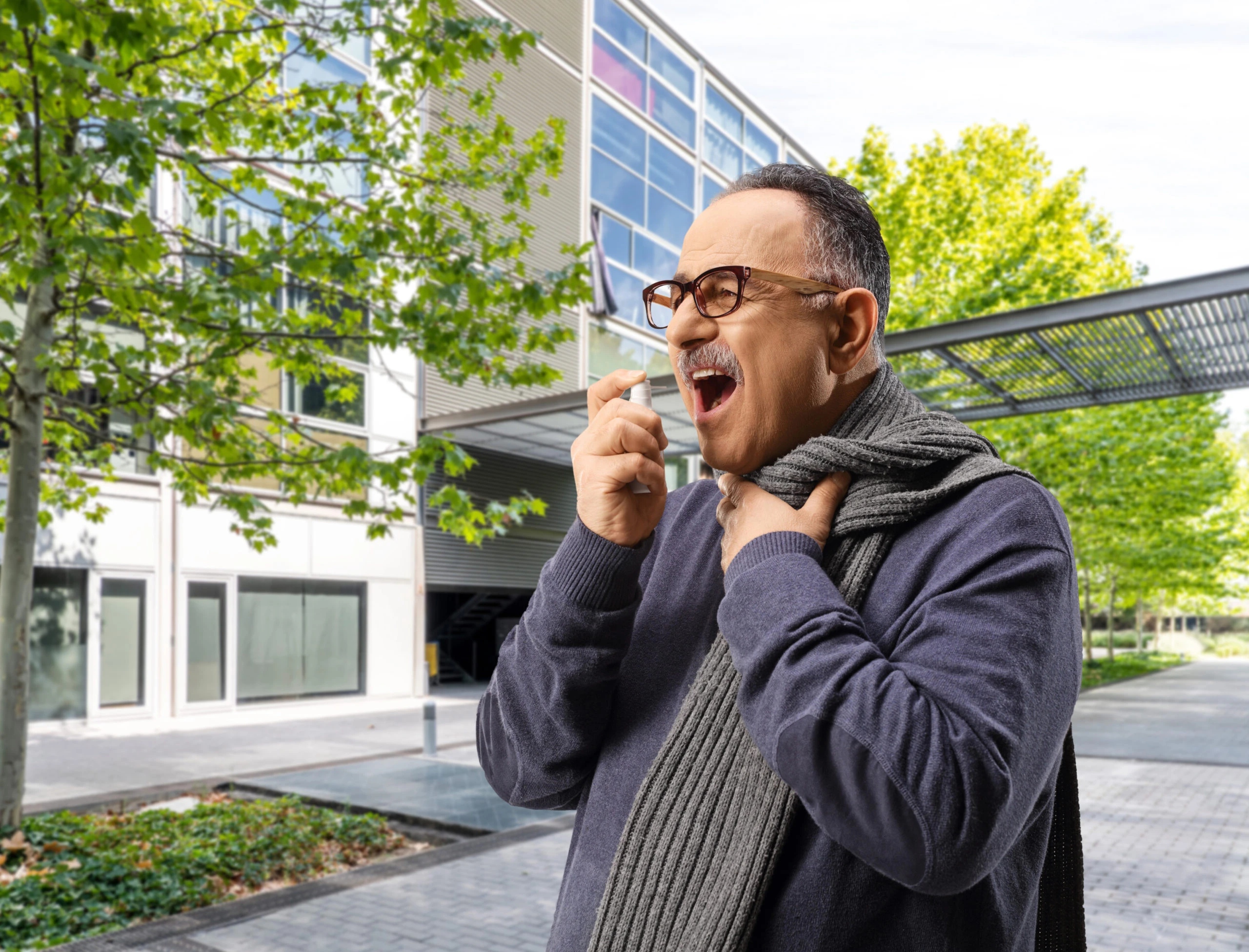 Older man in outdoor setting wearing a grey scarf and glasses dispensing liquid dropper of VMS product in his mouth.