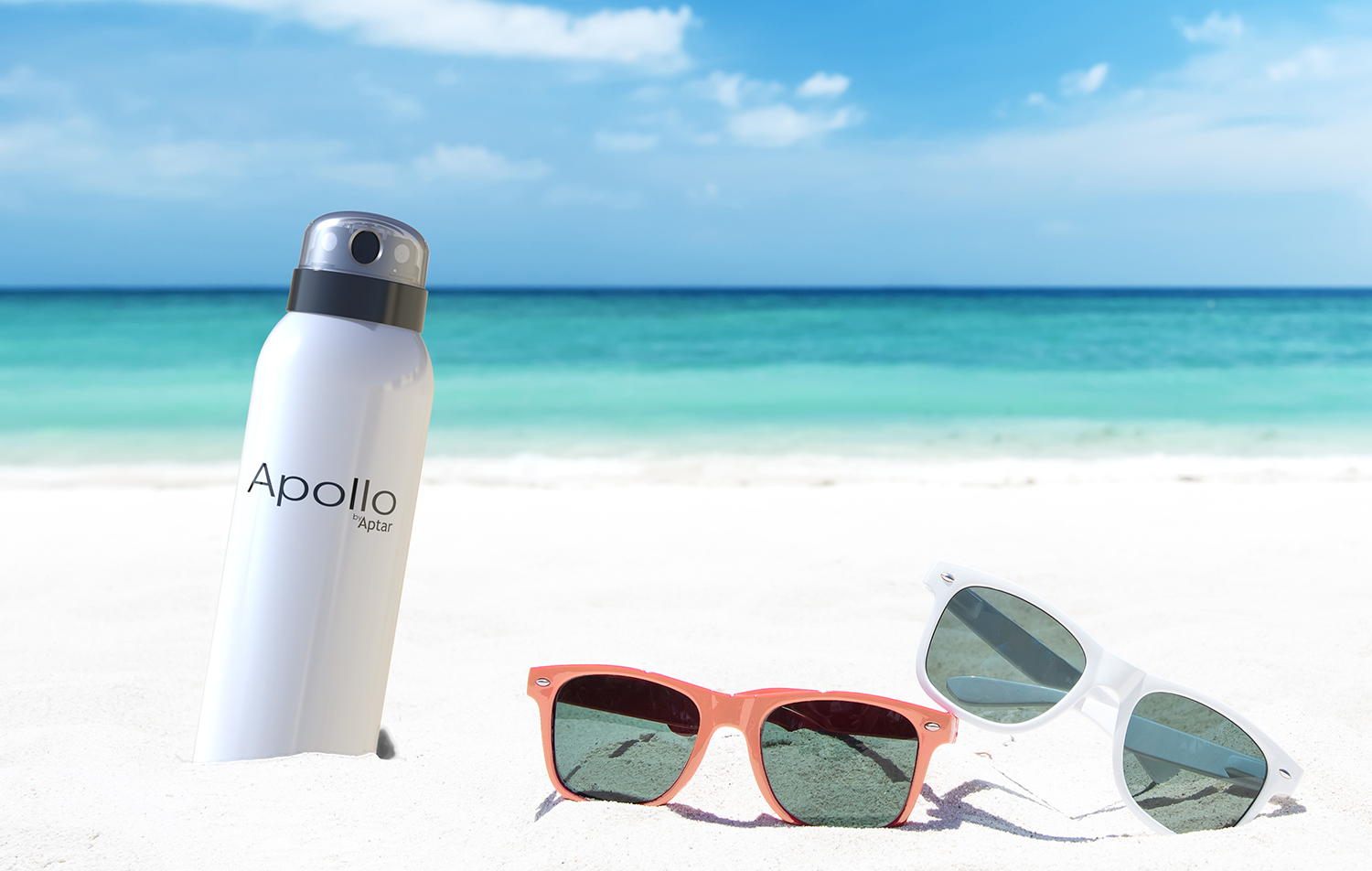 A bottle labeled Apollo, a dual-spray aerosol actuator, and two pairs of sunglasses rest on white sand, as turquoise ocean waves sparkle under the blue sky in the background.
