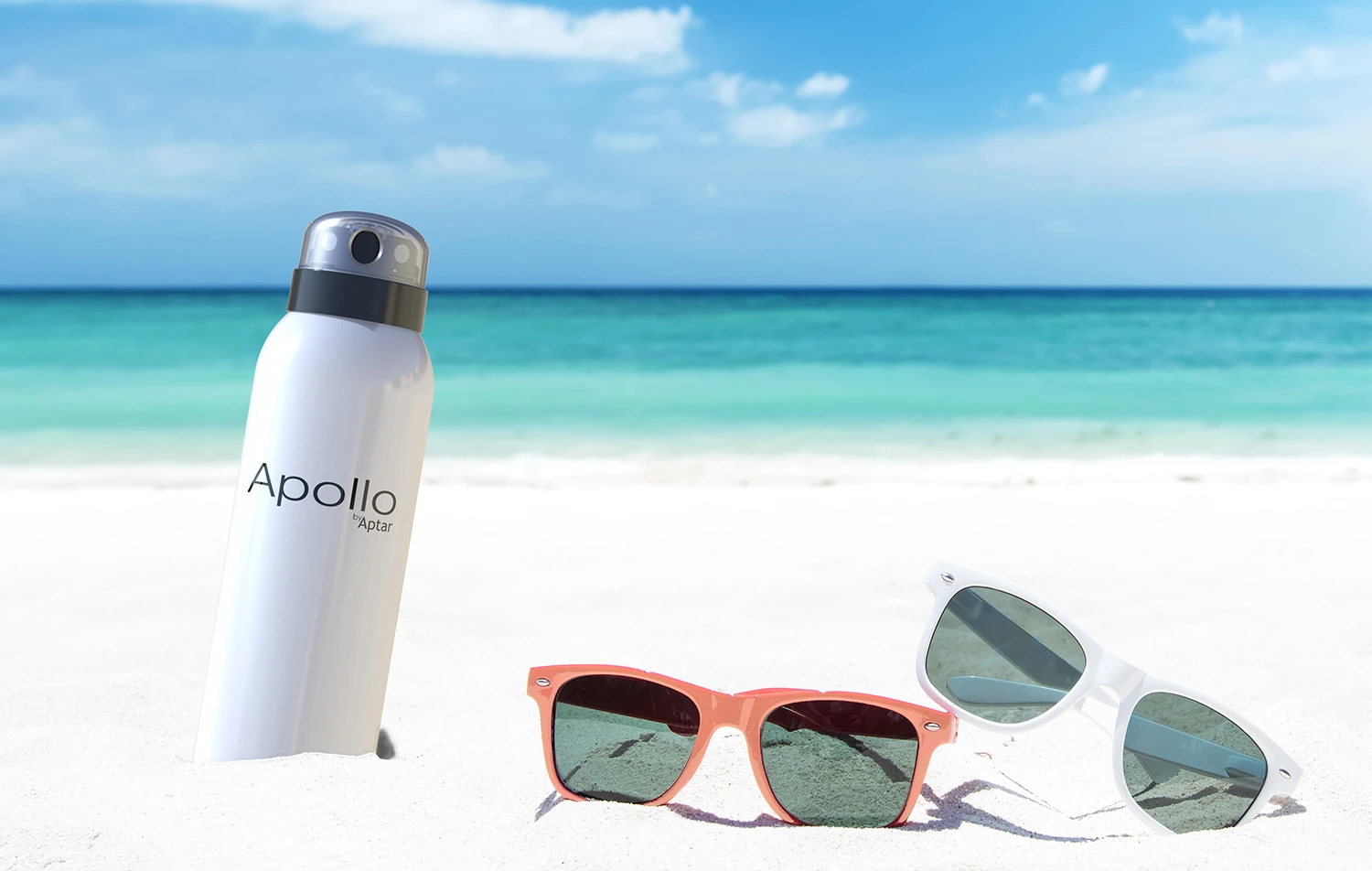 A bottle labeled Apollo, a dual-spray aerosol actuator, and two pairs of sunglasses rest on white sand, as turquoise ocean waves sparkle under the blue sky in the background.