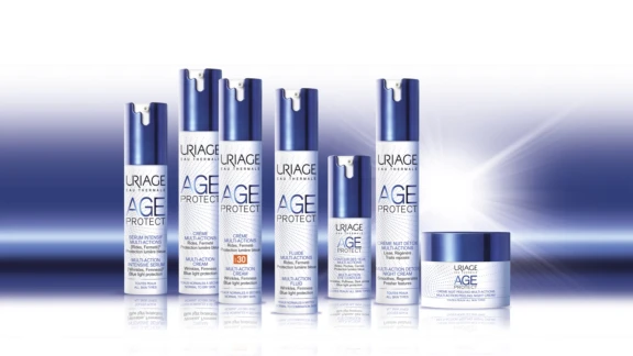A group of Uriage Age Protect skincare products is arranged on a reflective surface with a soft blue gradient background. The lineup includes several blue and white pump bottles and one jar, all featuring the premium airless packaging Auriga City. 