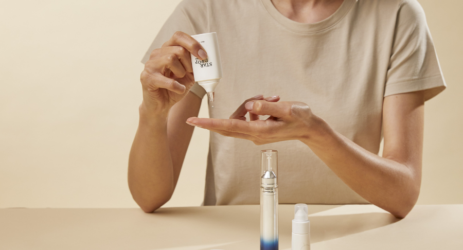 A person in a beige shirt uses drop-by-drop dispensing named Star Drop to squeeze skincare cream from a tube onto their fingers, with two other cosmetic bottles on the table in front of them, set against a neutral background.