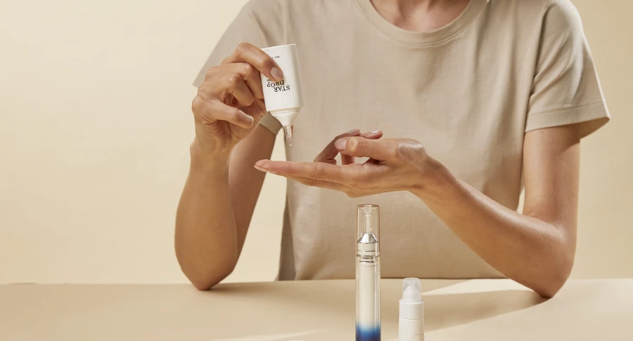 A person in a beige shirt uses drop-by-drop dispensing named Star Drop to squeeze skincare cream from a tube onto their fingers, with two other cosmetic bottles on the table in front of them, set against a neutral background.