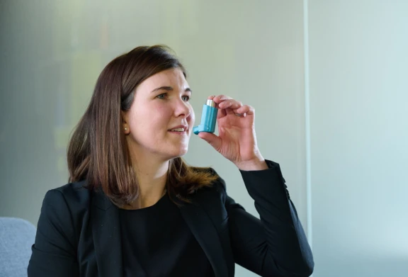 Woman with brown shoulder length hair in brown sweater holding blue inhaler containing Aptar inhaler metering valve.