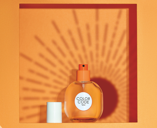An orange perfume bottle labeled COLOR CODE, featuring a plastic fragrance pump, sits in a square orange frame. The bottle casts a sunburst shadow on the orange background, while the white cap is placed beside it.