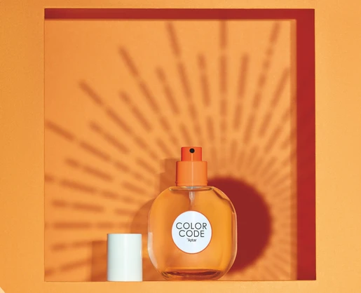 An orange perfume bottle labeled COLOR CODE, featuring a plastic fragrance pump, sits in a square orange frame. The bottle casts a sunburst shadow on the orange background, while the white cap is placed beside it.