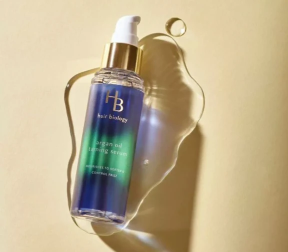 A bottle of Hair Biology argan oil taming serum with EuroMist Cardinal Lotion, a white low output cosmetic pump for high-viscosity formulas with a gold collar. The cap lies on a light surface.