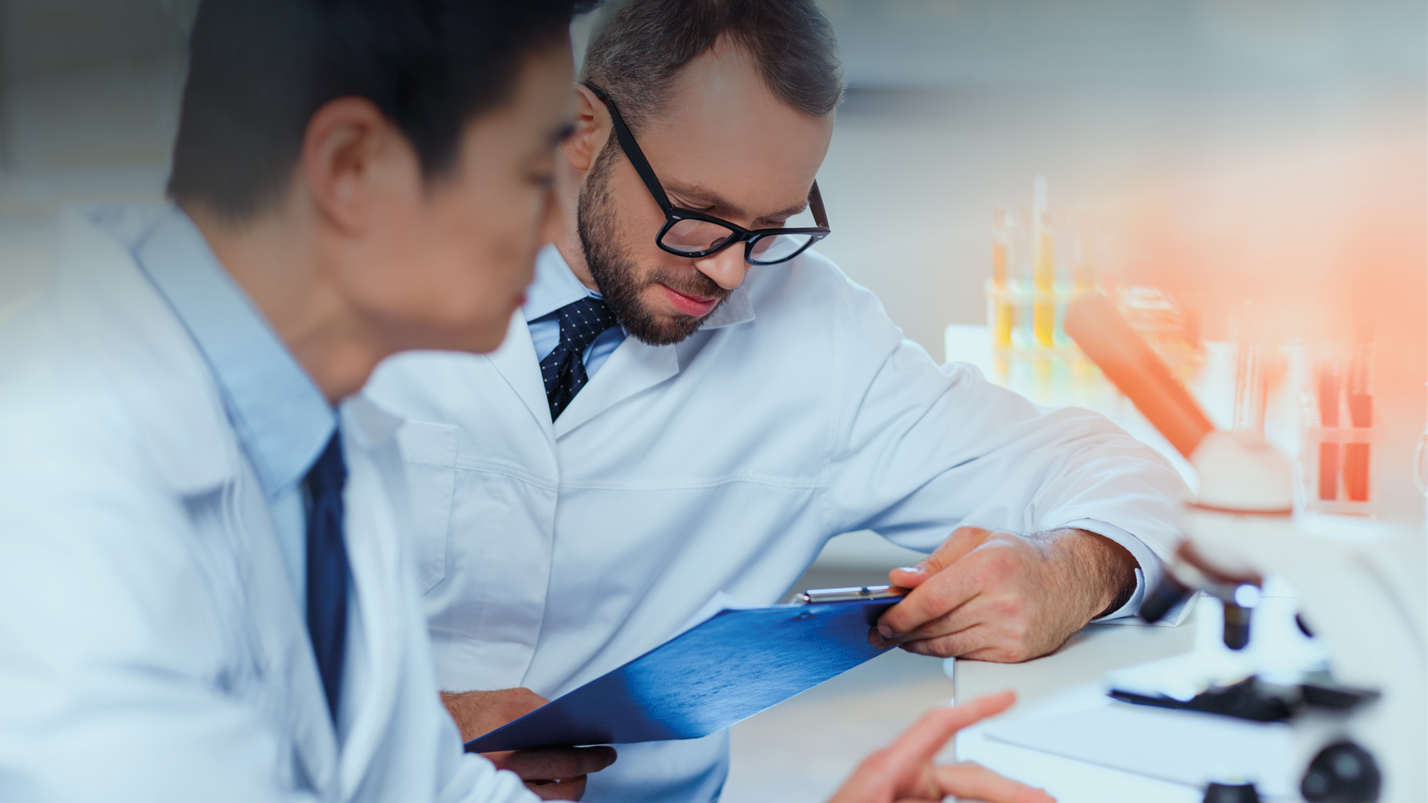 Two men sitting at laboratory benchtop in white lab coats review oral liquid product development data beside microscope.