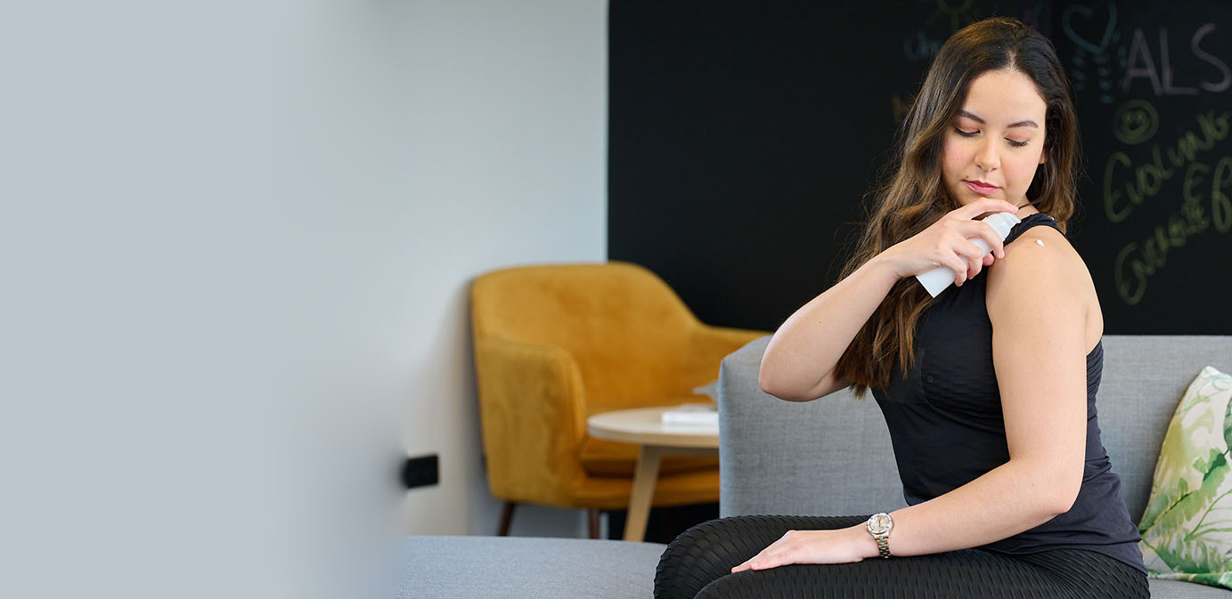 Woman in black sportswear seated on sofa applies dermal drug product to shoulder from Aptar Airless+ drug delivery system