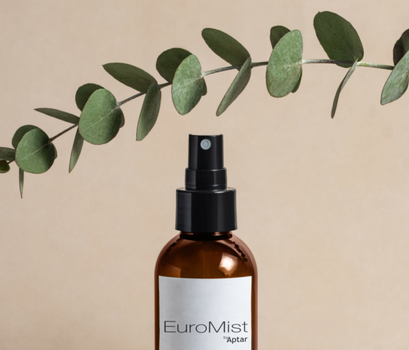 A brown spray bottle with a fine mist spray pump labeled EuroMist sits beneath a horizontal eucalyptus branch on a beige background.
