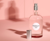 A clear glass perfume bottle labeled ESSENCIA SCREW by Aptar sits on a pink surface with its silver cap off, revealing the spray nozzle of a refillable fragrance pump. The background and shadows are also pink.