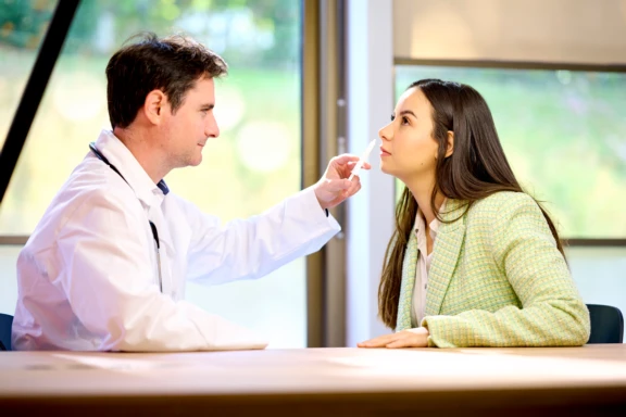 Male physician in white lab coat administers painless nasal vaccine to female patient in green jacket at doctor's office.