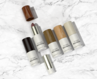Five lipsticks with wooden-textured lids in various shades, one with the lid off revealing a mauve lipstick, are arranged on a white marble background. The tubes feature ICONIC Woodacity and a unique Non-Guided Lipstick Mechanism for smooth application.