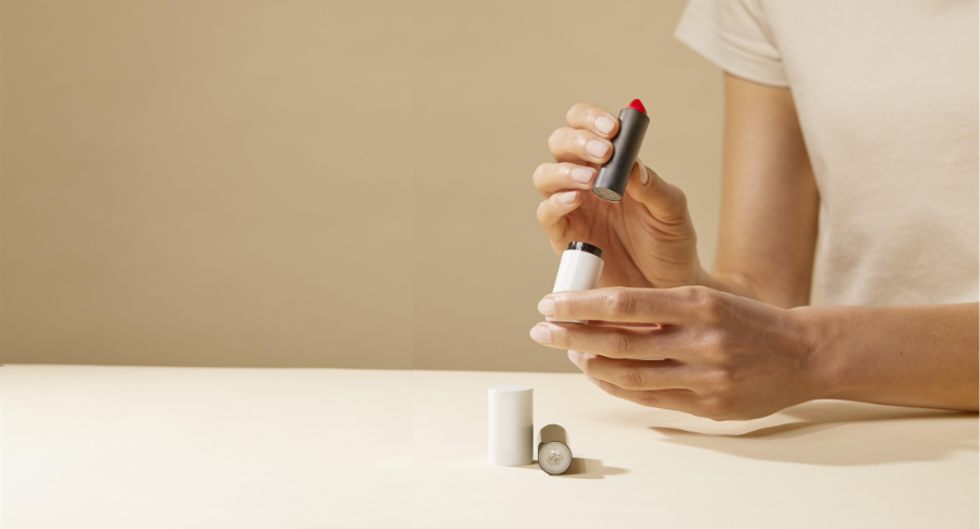 A person in a beige shirt holds an open tube of red lipstick above a light-colored table, showcasing sleek beauty packaging, with the lipstick cap and base resting on the surface.