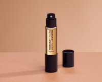 A small cylindrical perfume bottle with Refillable Mini Fragrance Packaging labeled NOMAD REFILL stands upright on a beige surface, its black cap beside it. The background features a gradient of light brown tones.