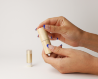 A person with blue French-tip nails holds a cylindrical bamboo container, while a gold tube featuring a Non-Guided Lipstick Mechanism labeled Iconic stands upright on a white surface in the background.