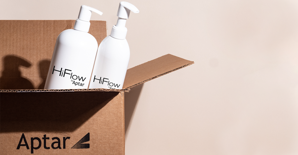 Two white bottles with e-commerce capable high-dose dispensing pumps labeled HiFlow Aptar stand inside an open brown cardboard box featuring the Aptar logo. The background is a plain light color.