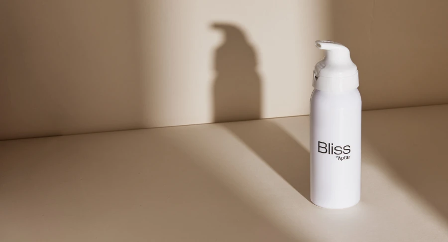 A white bottle with a dispenser pump labeled Bliss stands on a beige surface, its shadow cast elegantly on the wall a perfect example of luxury beauty dispensing.
