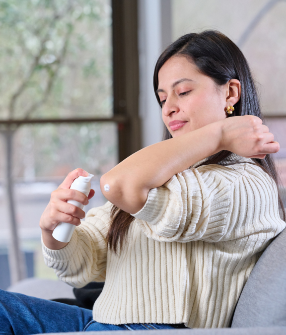 Woman with long brown hair seated in chair dispenses dermal cream on elbow from Aptar Airless+ drug delivery system