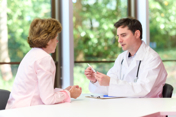 Male physician in white lab coat explains Aptar nasal vaccine device to female patient in doctor's office setting.