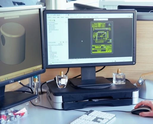 Two computer monitors on a desk display technical drawings; one shows a 3D object, the other a 2D schematic for custom primary packaging. A keyboard, perfume bottles, and a hand using a mouse are also visible.