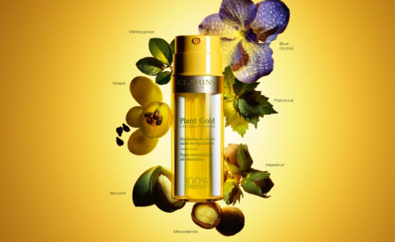 A yellow bottle of Clarins Plant Gold moisturizer, featuring a dual-chamber airless labeled Neomix design, is centered on a golden background and surrounded by labeled botanical ingredients: wintergreen, blue orchid, patchouli, hazelnut, macadamia, almonds, and grape.