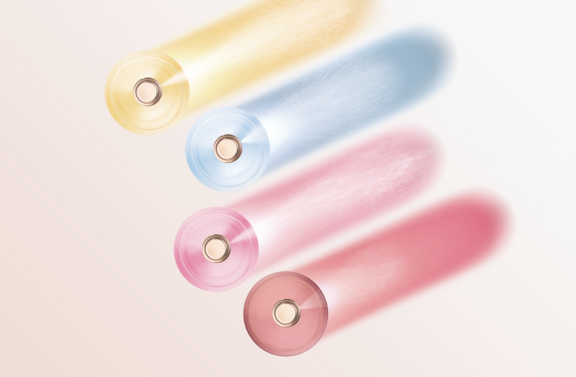 Four cylindrical objects, each in yellow, blue, pink, and red, roll forward like a classic prestige fragrance pump named Classic Inune in motion, leaving vibrant trails of color on a light background.