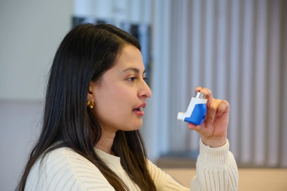Woman with long dark hair in white sweater holding blue Aptar Pharma DPI with integrated digital dose counter at home.