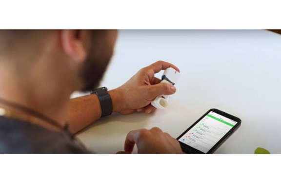 Bearded man holding Aptar Pharma digital health supported inhaler system while viewing smartphone.