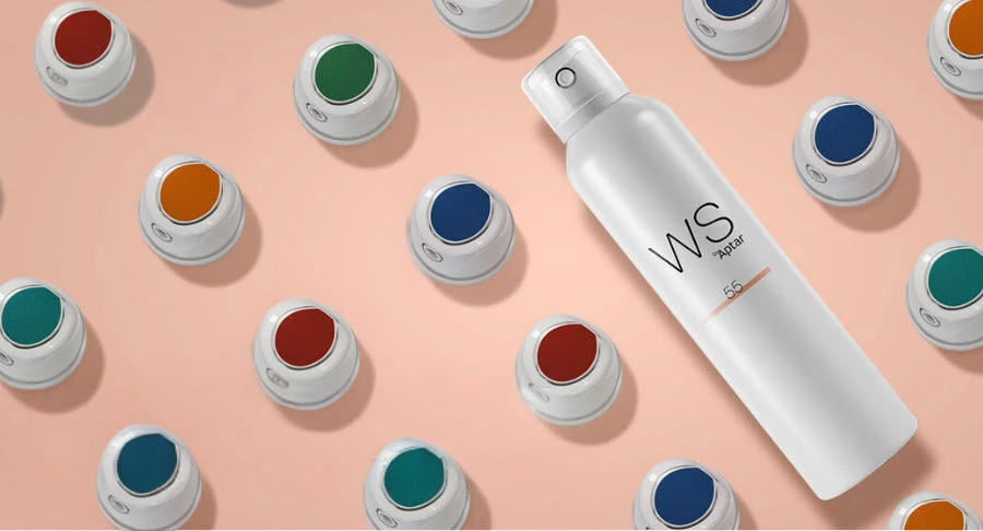 A white aerosol can with a white and silver Bicolor Aerosol Actuator labeled WS55 lies among rows of small white containers with colorful lids (green, blue, red, orange, and teal) on a peach-colored background.