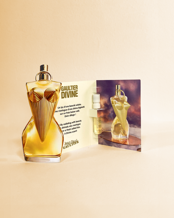 A uniquely shaped perfume bottle with a gold, bustier-style design stands next to an open fragrance sampling card featuring the same bottle, a small sample vial, and descriptive text.