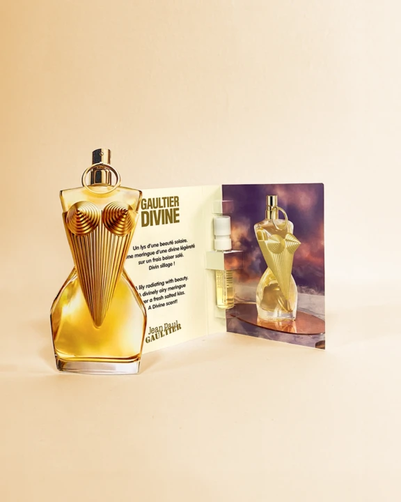 A uniquely shaped perfume bottle with a gold, bustier-style design stands next to an open fragrance sampling card featuring the same bottle, a small sample vial, and descriptive text.