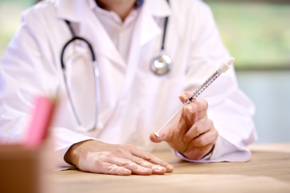 Doctor in white lab coat with stethoscope seated at wooden desk in office holding a clear Aptar nasal vaccine device.