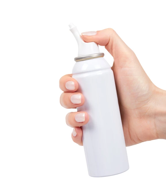 A hand holding and actuating a white bag-on-valve spray can. The user’s thumb presses down on the nozzle, demonstrating the ergonomic design of the actuator for controlled dispensing.