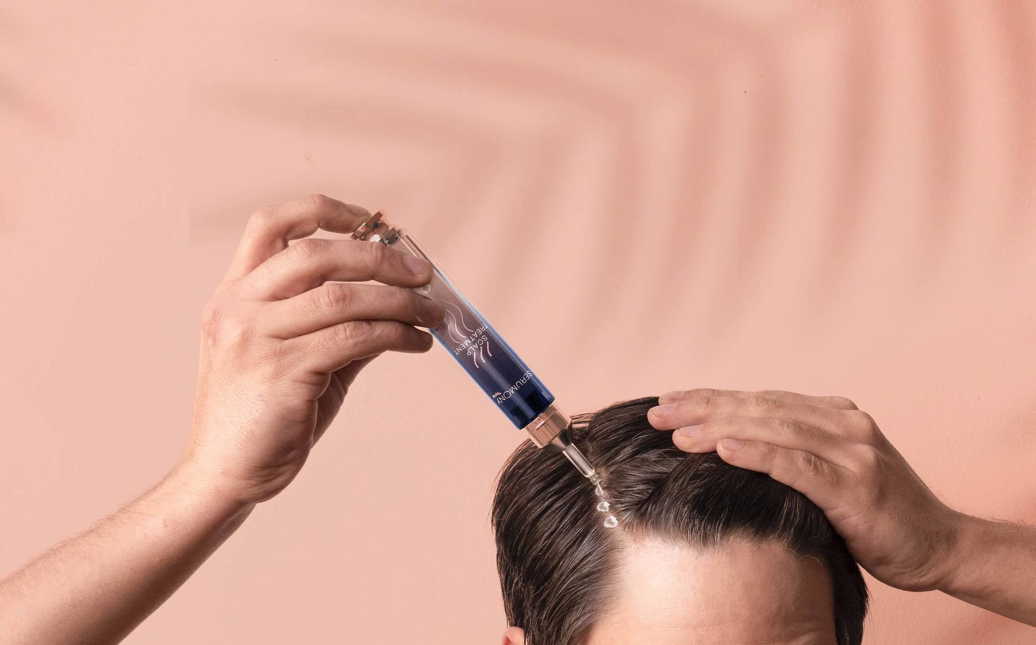 A person applies a blue liquid serum to their scalp using a dropper and sleek haircare packaging, with one hand parting their dark hair against a pale peach background.