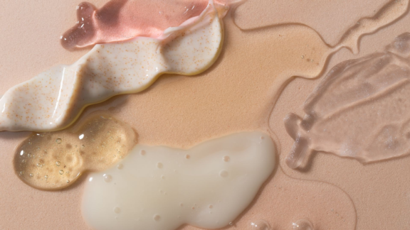 Various skincare or cosmetic creams, gels, and serums in different colors and textures are smeared and pooled on a smooth beige surface, highlighting both vibrant formulas and sleek haircare packaging.